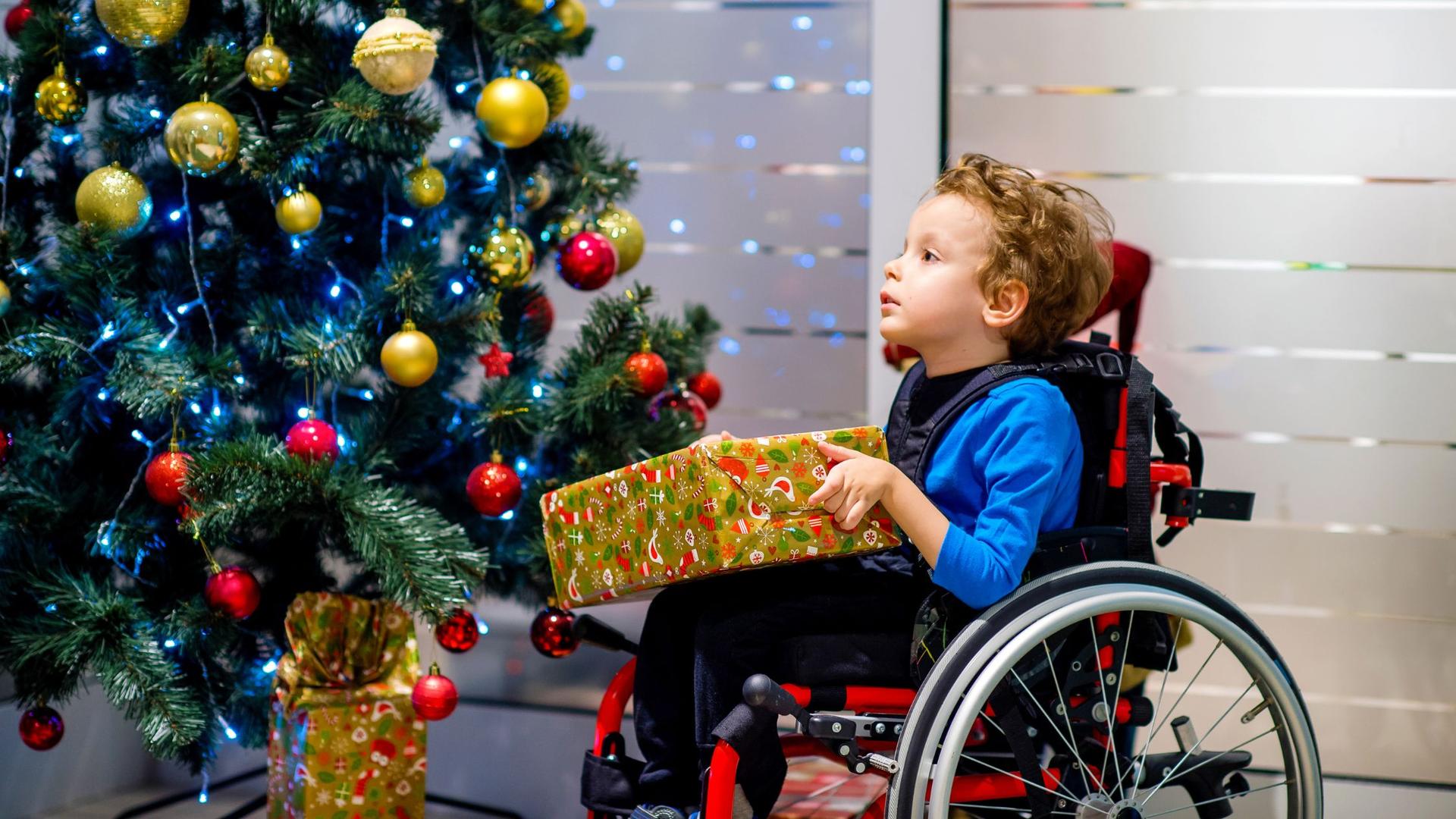 Child in a wheelchair holding a present, with a Christmas tree in the background.